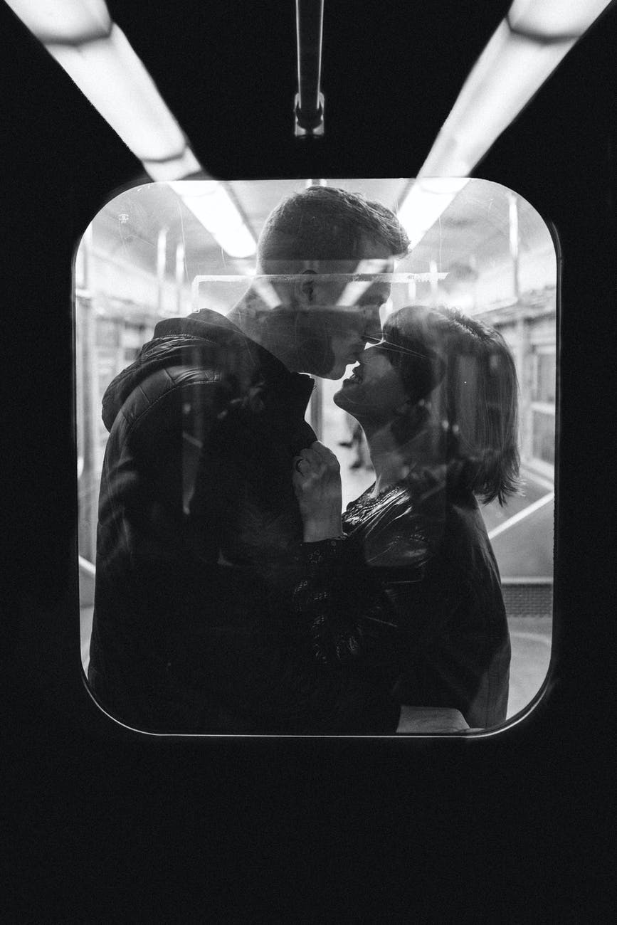 grayscale photo of man kissing woman s nose while standing inside a train