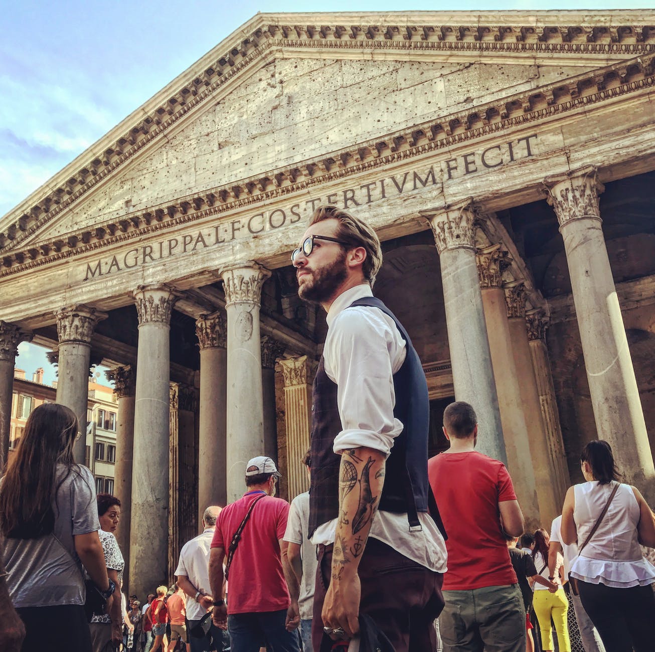 man in blue waistcoat and white dress shirt standing near magrippa lf costertivm fecit temple behind crowd of people